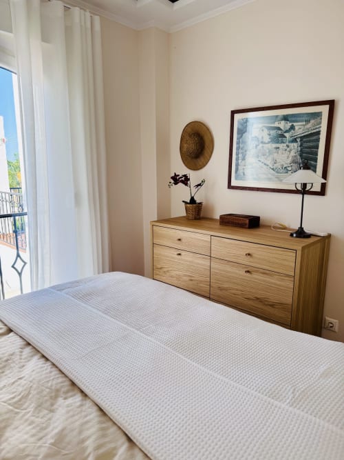 Dormitorio luminoso en villa de Nerja con cama blanca, cómoda de madera, cuadros enmarcados y balcón con vistas al mar