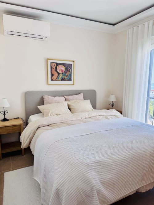 Dormitorio moderno en villa de Nerja con cama tapizada gris, ropa de cama crema, mesitas de madera y vistas al mar por puertas de cristal