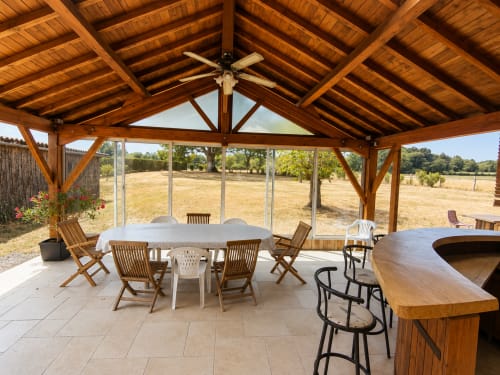 Covered patio with wooden beams at Domaine de Beau Site cottage, featuring dining table, bar seating, and countryside garden