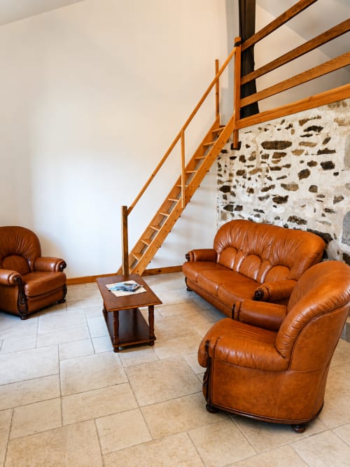 Cozy lounge area at La Longère cottage with leather armchairs, wooden staircase, and stone fireplace