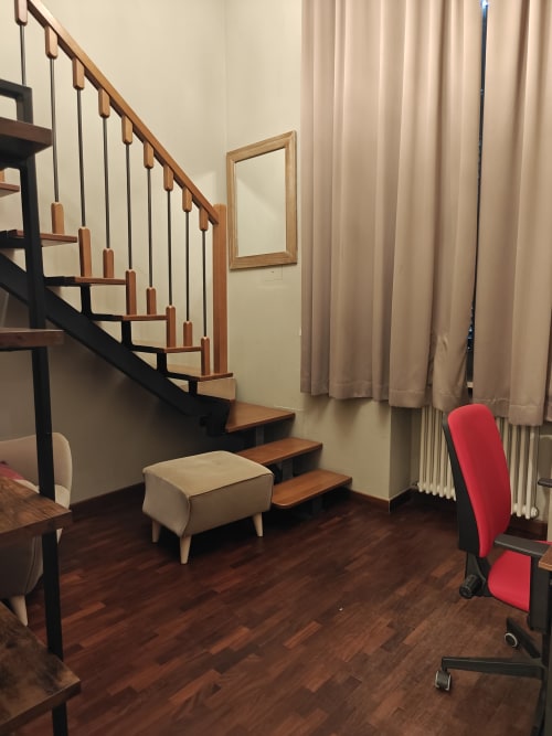 Moderno spazio abitativo con scala in legno, pouf beige, sedia rossa da ufficio e tende color crema a Cesena