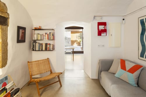Dormitorio minimalista moderno en el hotel Casa Merceditas en Corçà con silla de madera, sofá gris, estantería y puerta arqueada