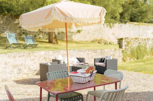 Espace de restauration en plein air du Hameau de Céline avec parasol crème, table colorée et cour en pierre dans les Landes
