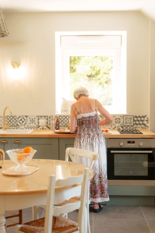 Femme âgée dans la cuisine du gîte Le Hameau de Céline, Landes-le-Gaulois, avec carrelage d'époque et vue sur le jardin