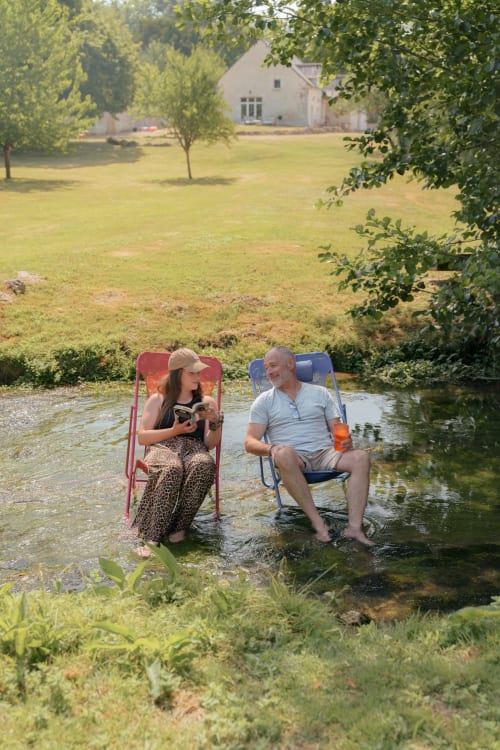 Deux hôtes se détendent dans des chaises de camping près d'un ruisseau au Hameau de Céline, un gîte à la campagne en Landes-le-Gaulois, France.
