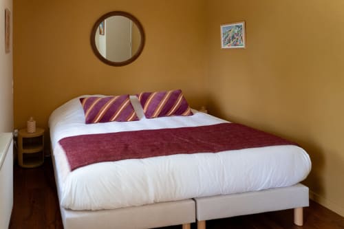 Chambre douillette aux Maisons Noisette avec coussins à rayures violettes, literie blanche, miroir rond et murs ocre chaleureux