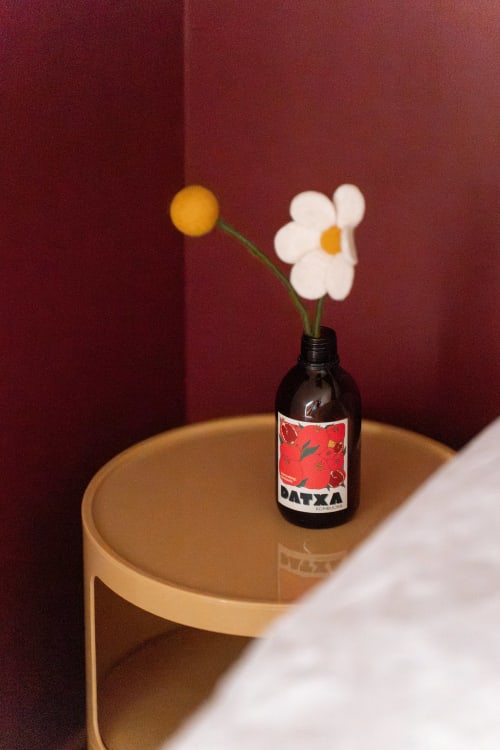 Bouteille de verre foncé à étiquette rouge sur table en bois devant mur bordeaux, avec fleurs blanches à cœur jaune aux Maisons