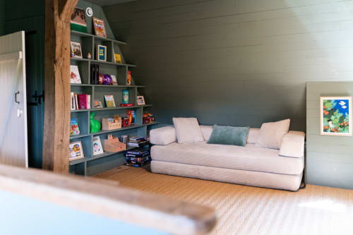 Coin lecture douillet chez Maisons Noisette avec canapé rose, étagères grises, livres et tableaux encadrés