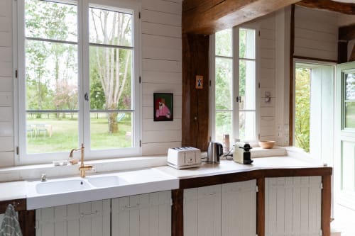 Cuisine lumineuse des Maisons Noisette avec plans de travail blancs, grandes fenêtres sur les jardins et poutres en bois rustique