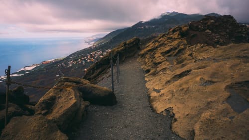 Sendero de senderismo volcánico con vista a la costa de La Palma, pueblos anidados en laderas bañadas por el sol y horizonte oceánico