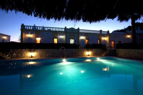 Piscina iluminada à noite com água turquesa, muro de pedra e elegante mansão na Herdade da Côrte em Tavira