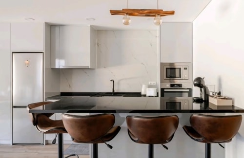 Cozinha moderna com ilha em granito preto, banquetas de couro marrom, armários brancos e luminária de madeira em Serração