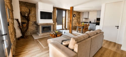 Sala de estar moderna da Pousada Serração em Rio Caldo com paredes de pedra, lareira, sofá bege, vigas de madeira e cozinha aberta.