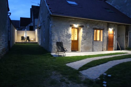 Cour du gîte Madiana 89 à Bellechaume au crépuscule, avec bâtisses en pierre aux fenêtres illuminées et coin détente