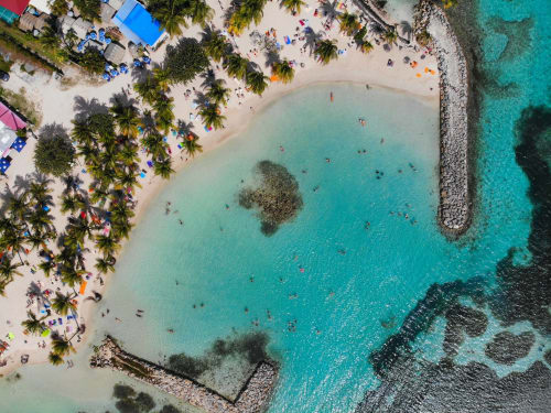 Vue aérienne des Villas Amboise en Guadeloupe avec plage aux eaux turquoise, palmiers et baigneurs sur sable blanc.