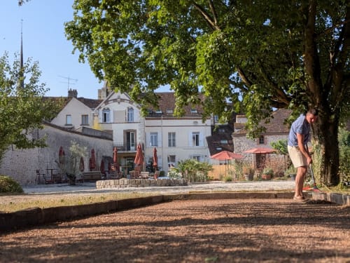 Homme jouant à la pétanque dans la cour du Domaine de La Poule Noire, entouré de bâtiments historiques français et d'arbres offrant de l'ombre