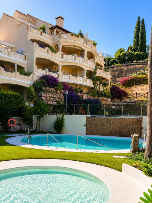 Área de piscina BH Marbella con piscinas de aguas turquesas, jardines exuberantes y edificio de apartamentos de estilo mediterráneo beige