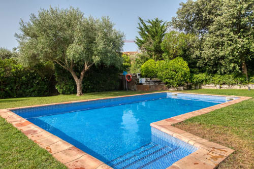 Piscina rectangular con agua azul rodeada de jardines cuidados, olivos y exuberante vegetación en la villa La Pastora