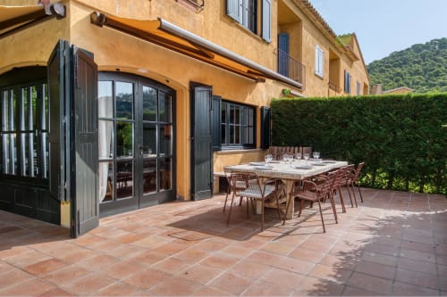 Patio de terracota con mesa de comedor y sillas en la villa La Pastora en Palafrugell, España, con puertas de marco negro y exuberante vegetación
