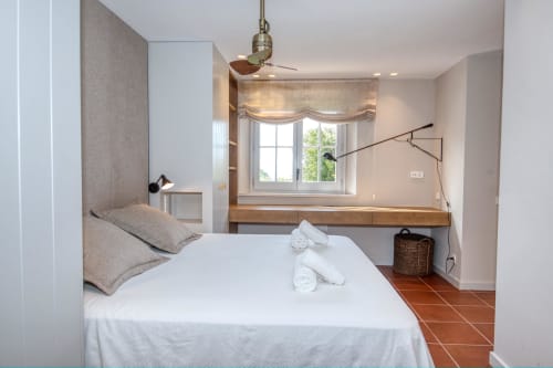 Dormitorio luminoso en villa Llafranc con ropa de cama blanca, escritorio de madera, ventilador de techo y suelos de terracota