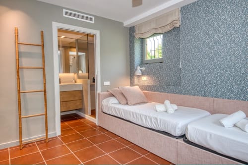 Elegante habitación doble con camas tapizadas en rosa, papel pintado con estampado azul y baño privado en la villa Llafranc.