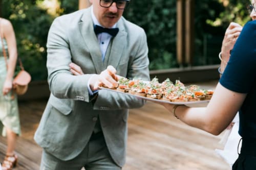 Man in gray blazer and bow tie serving appetizers at Ô Château hotel outdoor event in Hayange