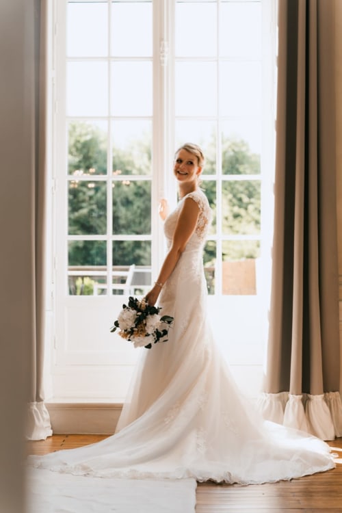 Bride in white lace wedding dress with bouquet at Ô Château hotel in Hayange, posed by windows
