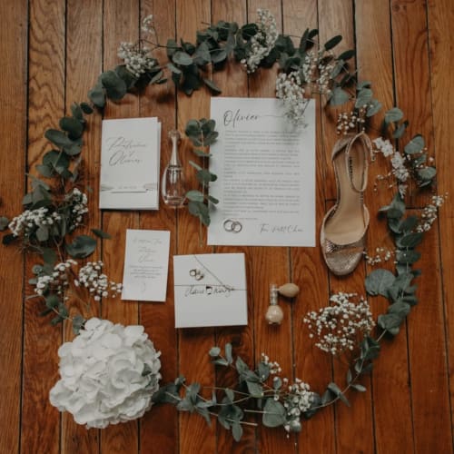 Wedding invitation flat lay for Ô Château in Hayange with eucalyptus garland, baby's breath, heels, and rings on wooden floor