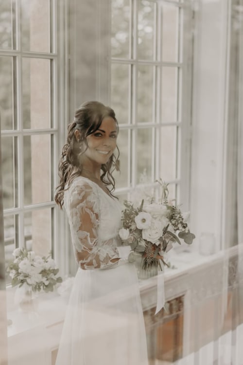 Bride in lace white dress with bouquet at Ô Château hotel window in Hayange