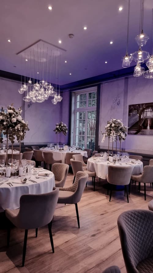 Elegant purple-lit banquet hall at Ô Château Hayange with round tables, white linens, upholstered chairs, crystal pendant