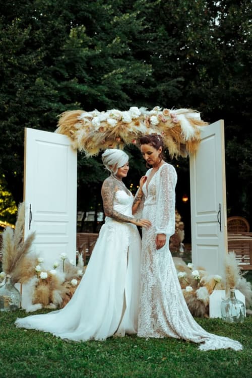 Two brides in white lace gowns at Ô Château wedding venue in Hayange, under ornate floral arch