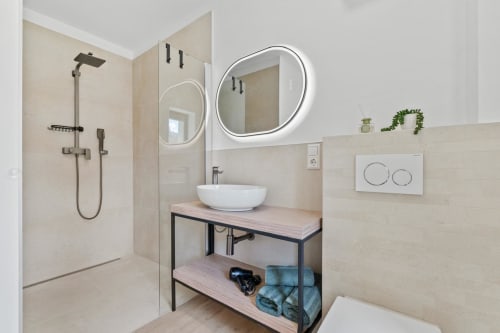 Salle de bains moderne chez Mattbnb Metz avec miroir rond lumineux, vasque blanche, meuble en bois et douche à l'italienne avec carrelage beige