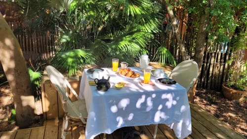 Petit-déjeuner au Lodge du Cap Ferret avec table, chaises, viennoiseries, jus de fruit et jardin tropical en arrière-plan.