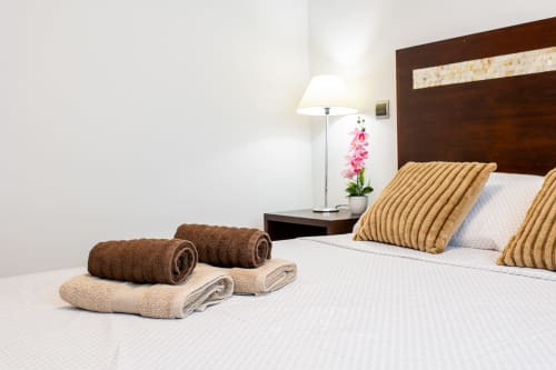 Bright bedroom at Haus Holiday Homes with towels on bed, gold pillows, dark wood headboard, and pink flowers on nightstand
