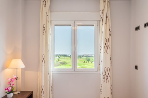 Bright apartment room with cream curtains, table lamp with pink flowers, and double doors opening to golf course views at