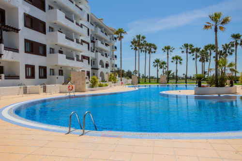 Large curved swimming pool with palm trees and white apartment building at Haus Holiday Homes in San Pedro del Pinatar, Spain