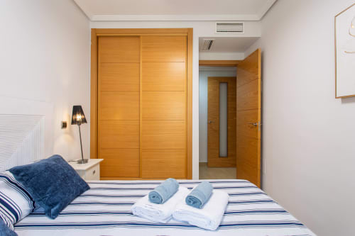 Bedroom with striped bedding, blue pillows, rolled towels, wooden wardrobe, and bedside lamp at Haus Holiday Homes, San Pedro