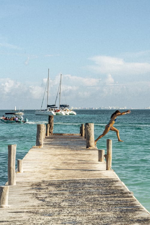Man diving off wooden pier in Isla Mujeres with sailboats and turquoise water visible