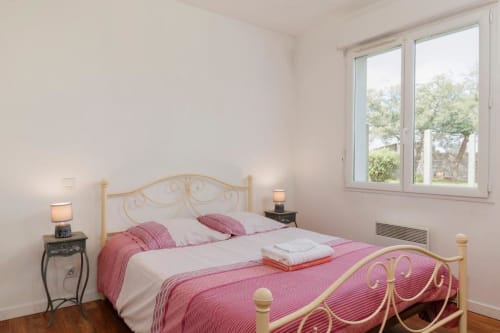 Chambre lumineuse du gîte Les Petits Toits à Libourne avec lit en métal, draps roses, fenêtres sur jardin et commode