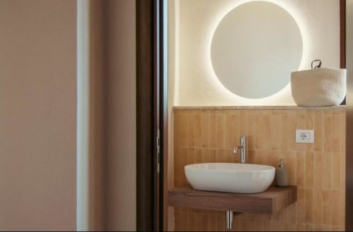 Bagno moderno della suite Rosmarino al Baglio Capo con specchio circolare illuminato, lavabo bianco su bancone in legno