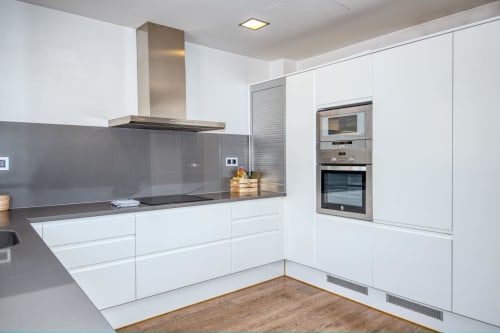 Modern white kitchen in Calella apartment with stainless steel appliances, gray backsplash, and wooden flooring