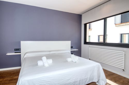 Modern bedroom in Calella apartment with purple accent wall, white bed, and large windows overlooking the town