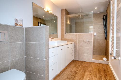 Modern bathroom in Calella apartment with walk-in shower, white vanity, wooden flooring, and gray tile walls