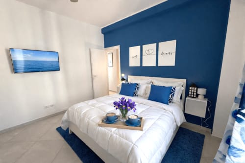 Camera moderna degli Appartamenti Bari con parete azzurra, biancheria bianca, fiori viola, TV e vista sulla città di Bari