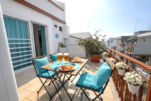 Terrazza soleggiata degli Appartamenti Bari con sedute turchesi, cibo, bevande, fiori e vista sulla città
