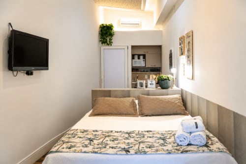 Quarto aconchegante Loft ROMA no CSS Maringá com TV de parede, claraboia, plantas e toalhas dobradas na cama