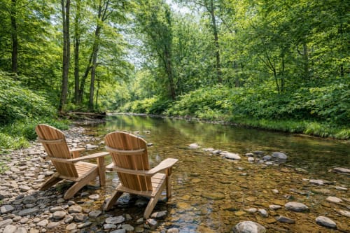 Two wooden Adirondack chairs on rocky shore facing calm creek in lush forest, Malkita Terra villa outdoor setting