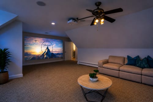 Modern home theater room with tan sectional sofa, round wood coffee table, ceiling fan, and large projection screen