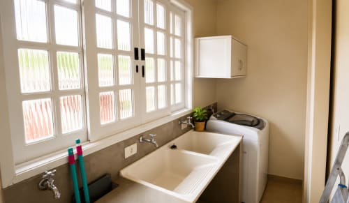 Lavanderia iluminada do apartamento CSS em Maringá com pia dupla, máquina de lavar, vista pela janela e armários