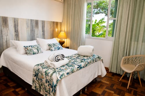 Quarto luminoso com roupa de cama branca, almofadas estampadas, cabeceira de madeira, luminária lateral e vista para o jardim no apartamento de Maringá